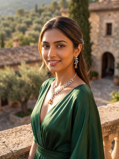 Woman in a green dress with jewelry standing in an outdoor setting with stone buildings and trees.